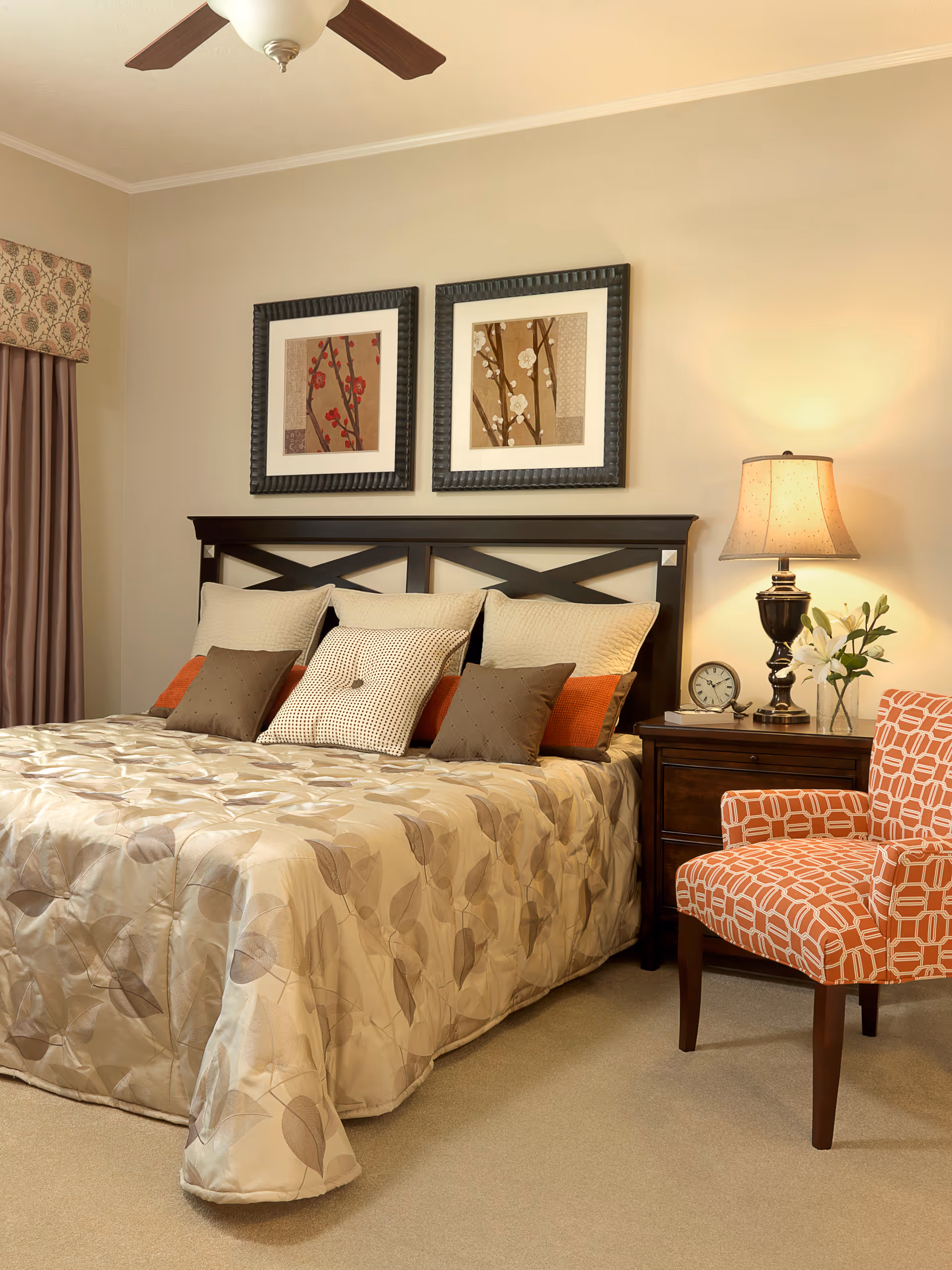 A neatly made bed with a beige and brown leaf-patterned comforter and multiple pillows in beige, brown, and orange shades. Above the bed are two framed floral artworks. To the right of the bed is a wooden nightstand with a table lamp, a clock, a book, and a vase with white flowers. An orange patterned armchair is positioned next to the nightstand. The room has beige walls and carpet, with a ceiling fan visible at the top.