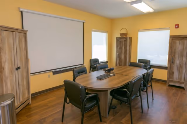Small meeting room with an oval wooden table surrounded by chairs, a wall-mounted projection screen, and wooden cabinets.