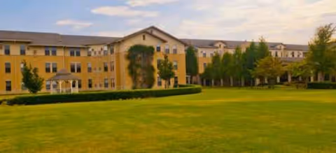A beige, multi-story senior living facility with a green lawn and trees in front.