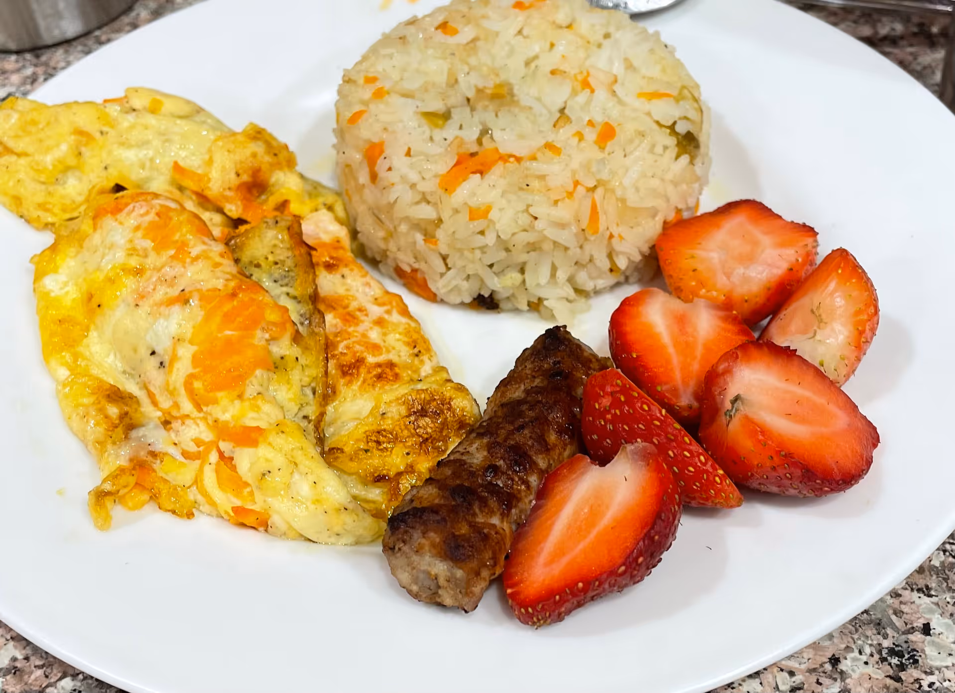 A white plate with a serving of rice, an omelette, a sausage link, and sliced strawberries on a countertop.
