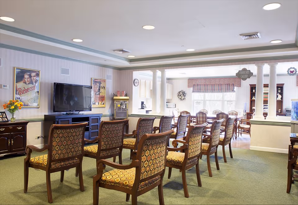 Bright senior living community room with rows of patterned wooden chairs facing a television and a dining area beyond.