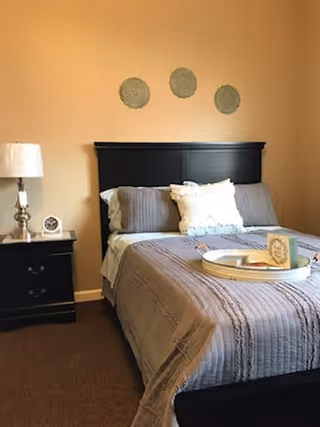A neatly made bed with a black headboard and gray bedding, featuring a white decorative pillow. A round tray with a card and some items is placed on the bed. To the left of the bed is a black nightstand with a white lamp and a small clock. Three decorative round wall hangings are displayed above the headboard on a beige wall.