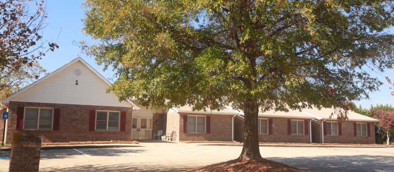 Single-story brick senior living facility exterior with a large tree, parking lot, and entrance.