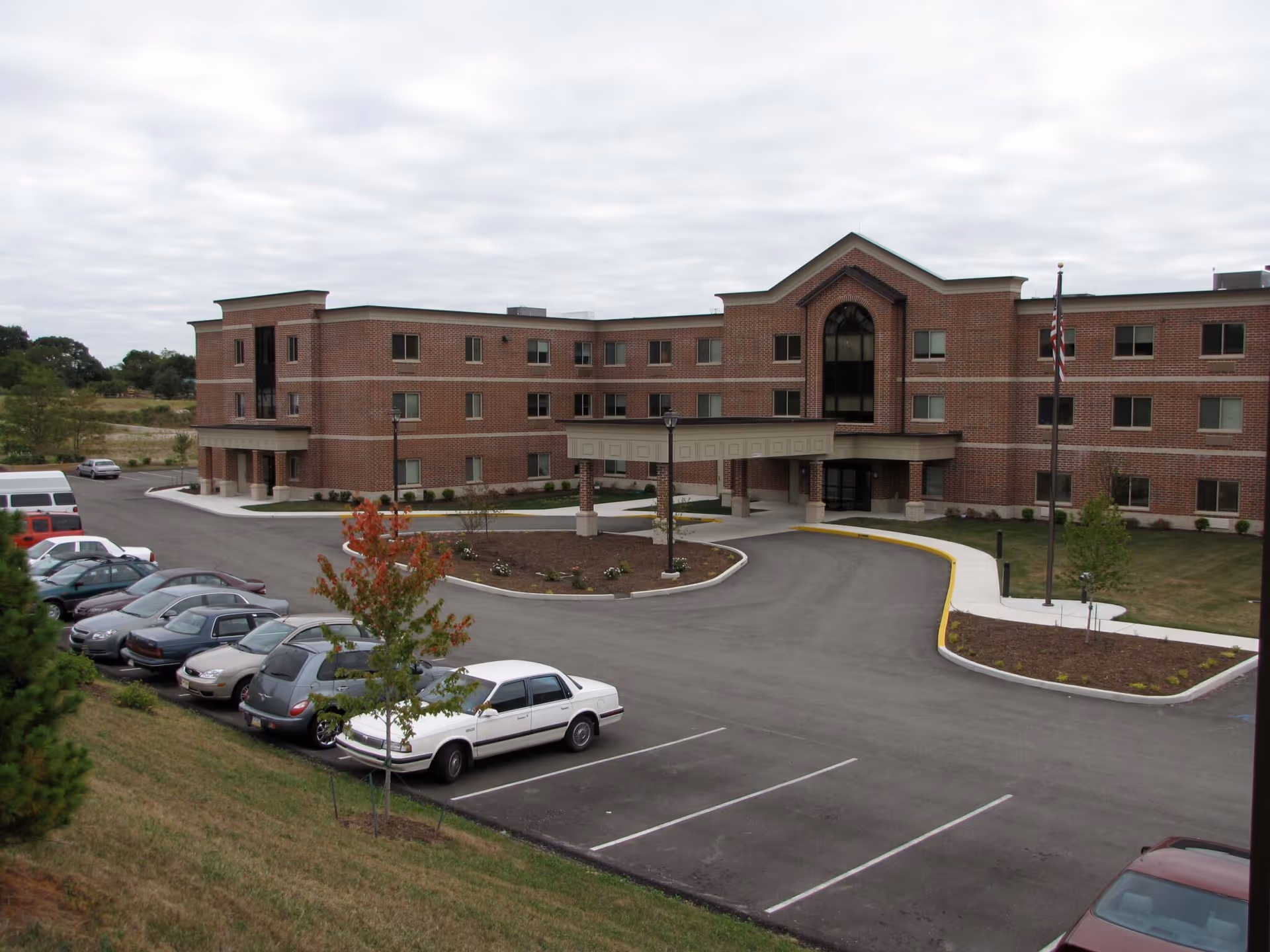 Exterior view of a three-story brick building with a covered entrance and a circular driveway. Several cars are parked in the parking lot in front of the building, and there is a flagpole with an American flag near the entrance. The sky is overcast.