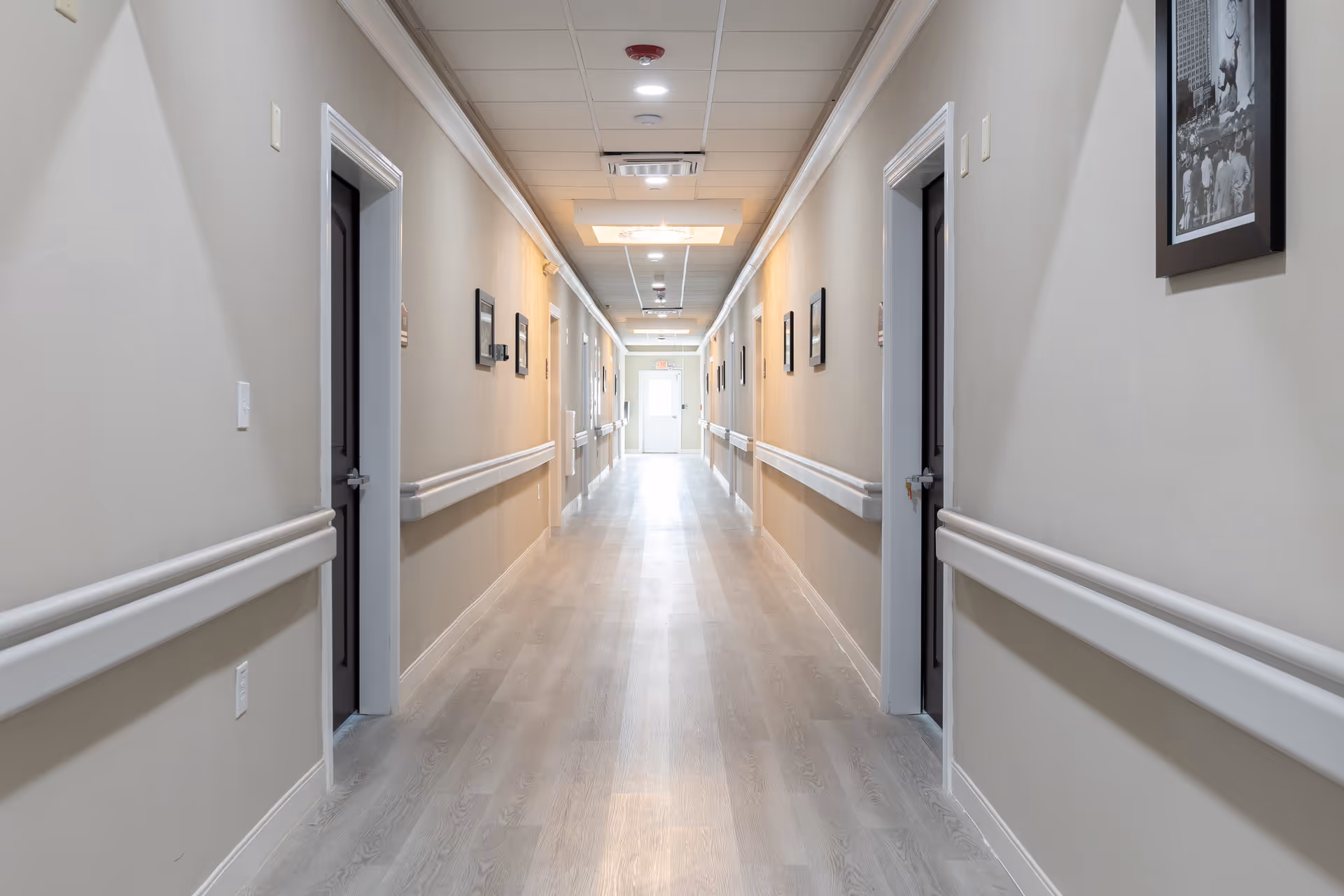A long, well-lit hallway in an assisted living facility with beige walls, light wood flooring, and multiple doors on both sides. Handrails run along the walls, and framed black-and-white photographs are hung at regular intervals. The ceiling has recessed lighting and a decorative light fixture near the center. A door with a window is visible at the far end of the hallway.