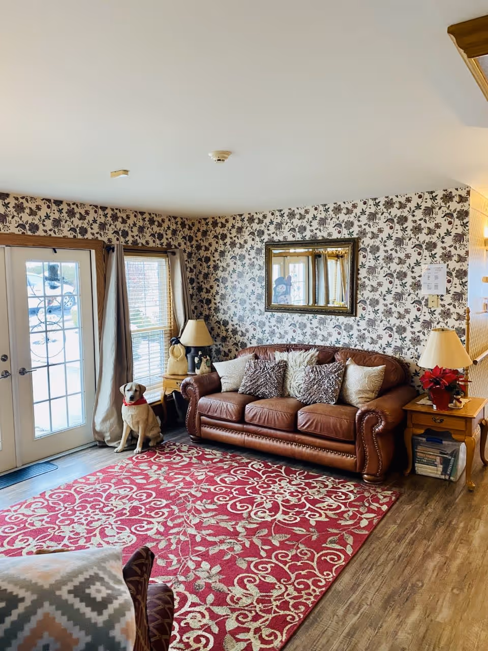 A cozy living room with a brown leather sofa adorned with multiple decorative pillows, a red patterned area rug on wooden flooring, floral wallpaper, two wooden side tables each with a lamp, a large mirror on the wall, and a dog sitting near the glass double doors with curtains.