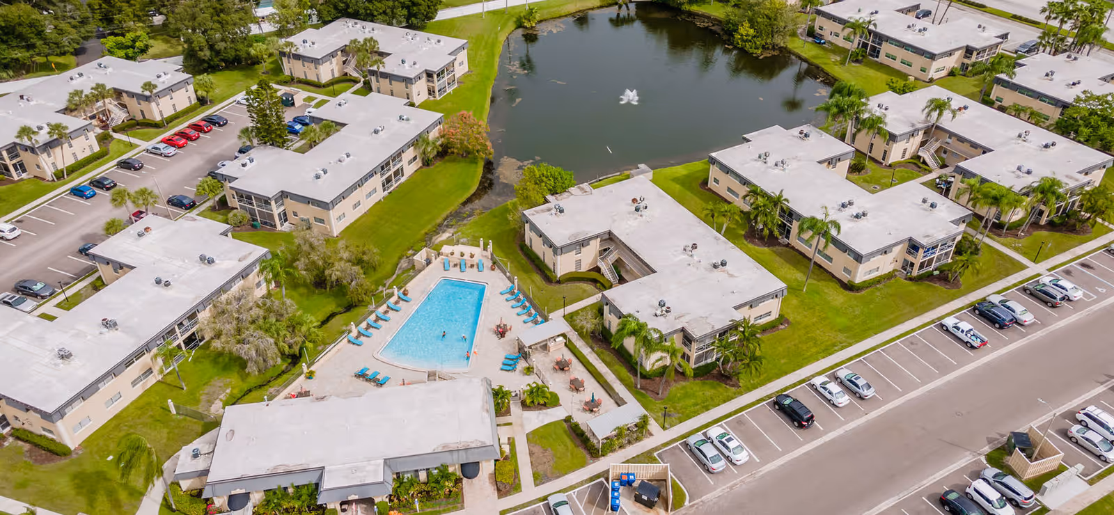 Aerial view of a residential complex with multiple low-rise buildings surrounding a central swimming pool and a pond.