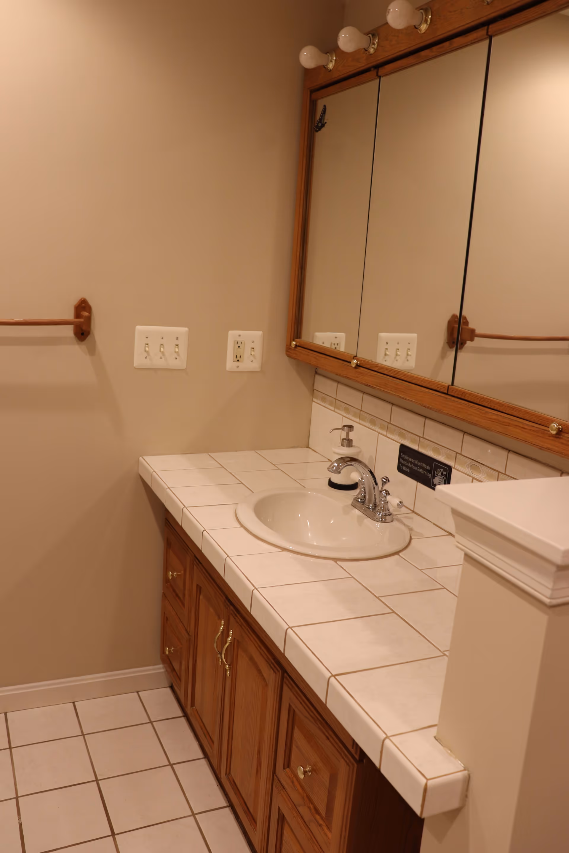 Bathroom vanity with a white tiled countertop and an inset sink. Above the sink is a large mirror with a wooden frame and three light bulbs mounted on top. There is a soap dispenser on the countertop and a towel bar mounted on the beige wall to the left. The floor is tiled with white tiles and brown grout.