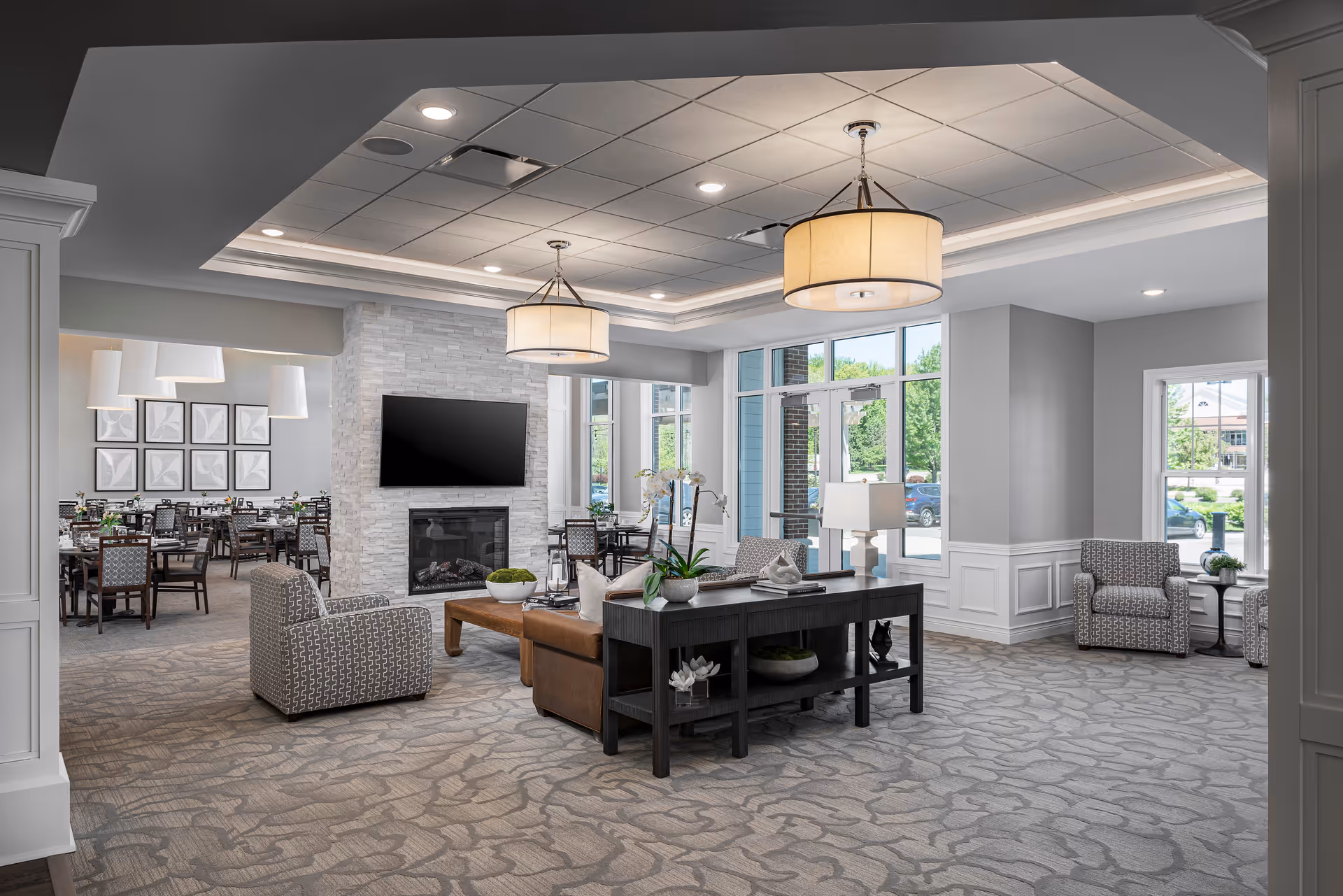 A spacious and well-lit common area in a senior living facility featuring a seating area with patterned armchairs, a brown leather sofa, a wooden coffee table, and a console table with decorative plants. A modern stone fireplace with a mounted flat-screen TV is centrally located. Large windows and glass doors allow natural light to fill the room. In the background, a dining area with multiple tables and chairs is visible, illuminated by pendant lights.
