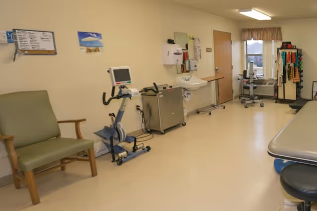 A rehabilitation room with exercise equipment including a stationary bike and resistance bands, a green armchair, a sink with a soap dispenser, a whiteboard on the wall, and a treatment table. There is a window with a valance and a door in the background.
