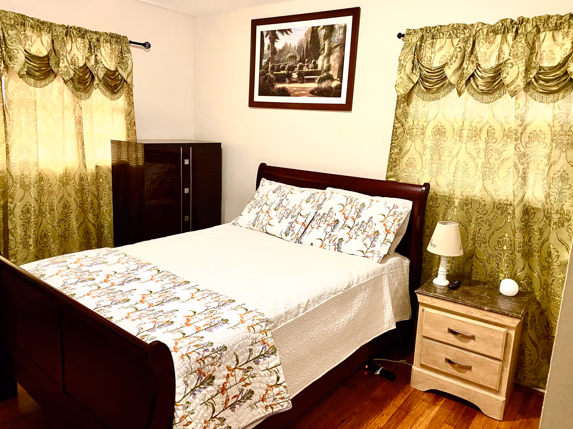 A cozy bedroom with a wooden bed frame, a white bedspread with floral patterns, matching pillows, two windows with ornate green curtains, a wooden dresser in the corner, a nightstand with a lamp and a small decorative item, and a framed landscape painting on the wall.