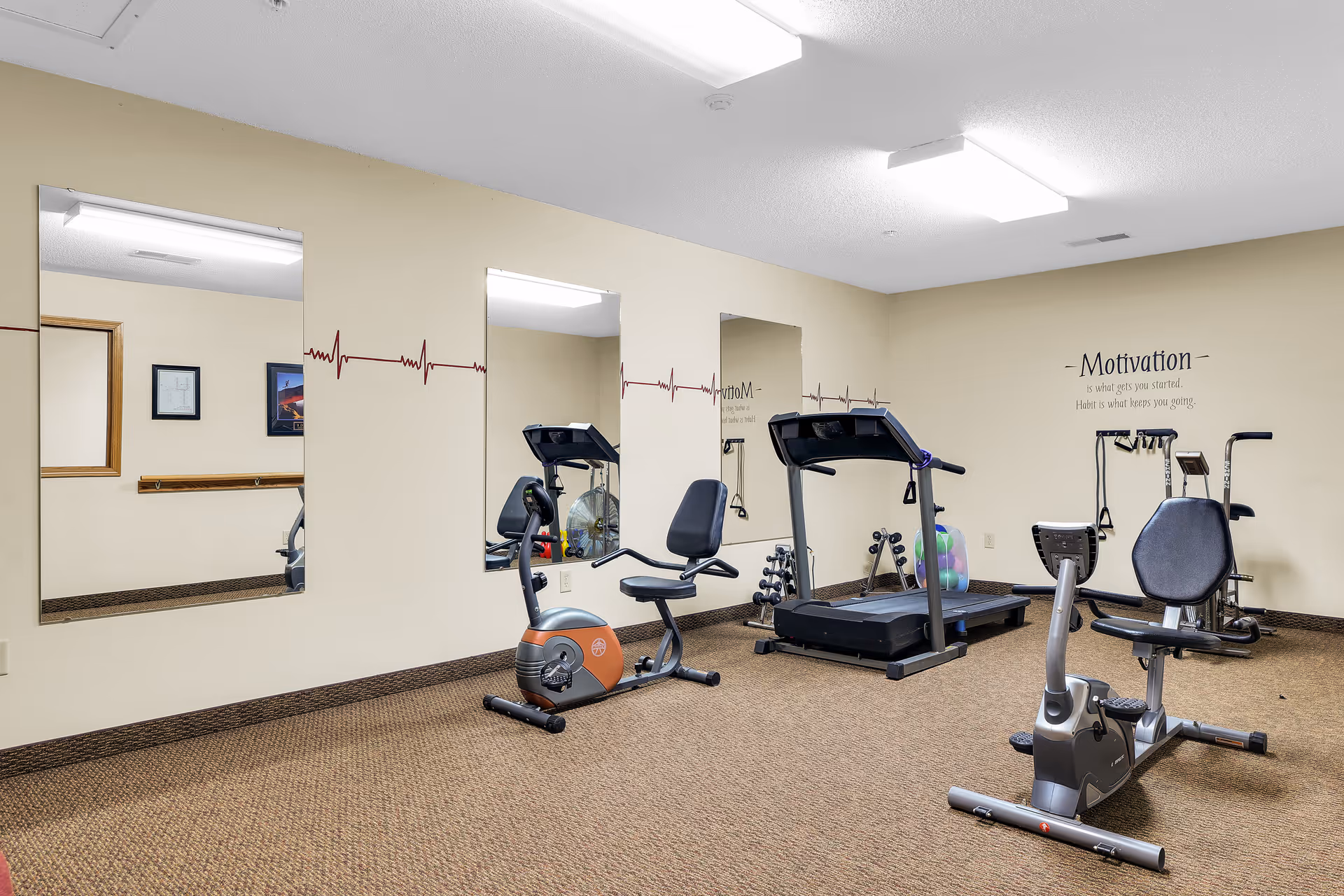 A small fitness room with exercise equipment including two recumbent stationary bikes, a treadmill, and a multi-exercise machine. The walls are beige with three large mirrors and a heartbeat line decal running horizontally. There is a motivational quote on the wall that reads, 'Motivation is what gets you started. Habit is what keeps you going.' The floor is carpeted in brown.