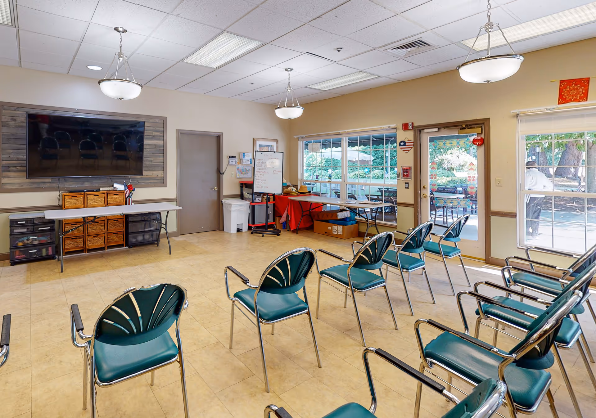 A well-lit room with multiple green chairs arranged in rows facing a large flat-screen TV mounted on the wall. There are tables along the walls with storage baskets and a whiteboard. Large windows and a glass door provide a view of an outdoor patio area with trees and seating.