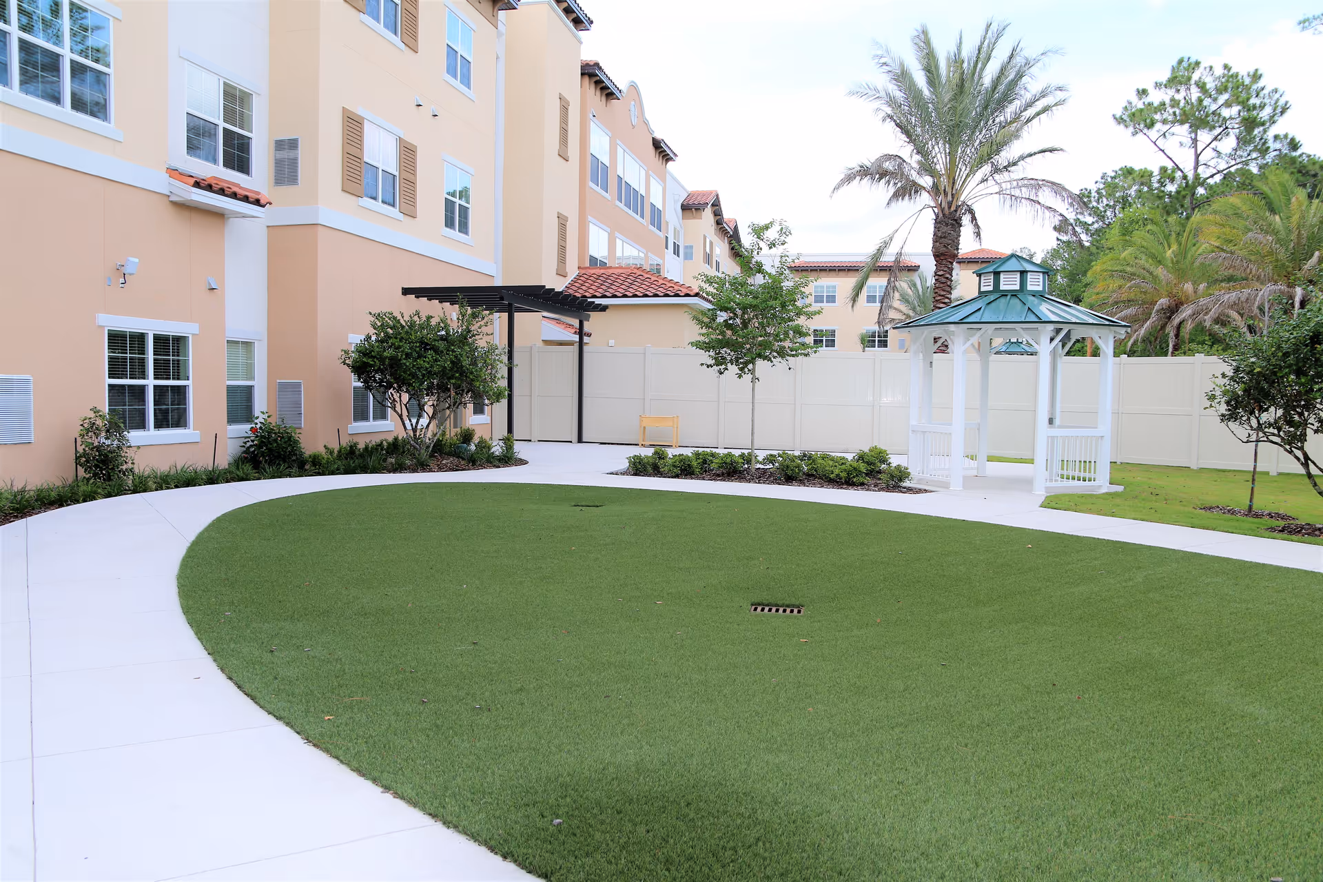 Outdoor courtyard with a green lawn, white gazebo, palm trees and a beige senior living building in the background.