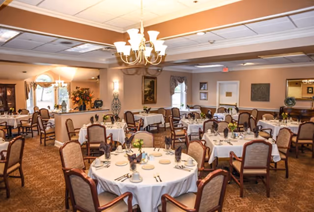 A well-lit dining room with multiple round and rectangular tables covered with white tablecloths, each set with plates, cups, silverware, and folded napkins. The room has beige walls, carpeted floors, and several chairs around each table. There are decorative elements including framed paintings, floral arrangements, and a chandelier hanging from the ceiling.