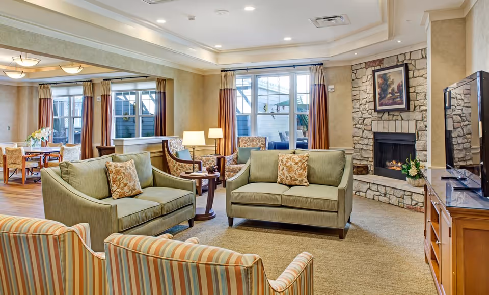 A bright communal living room with sofas, armchairs, a stone fireplace, a TV, and a dining area visible in the background.