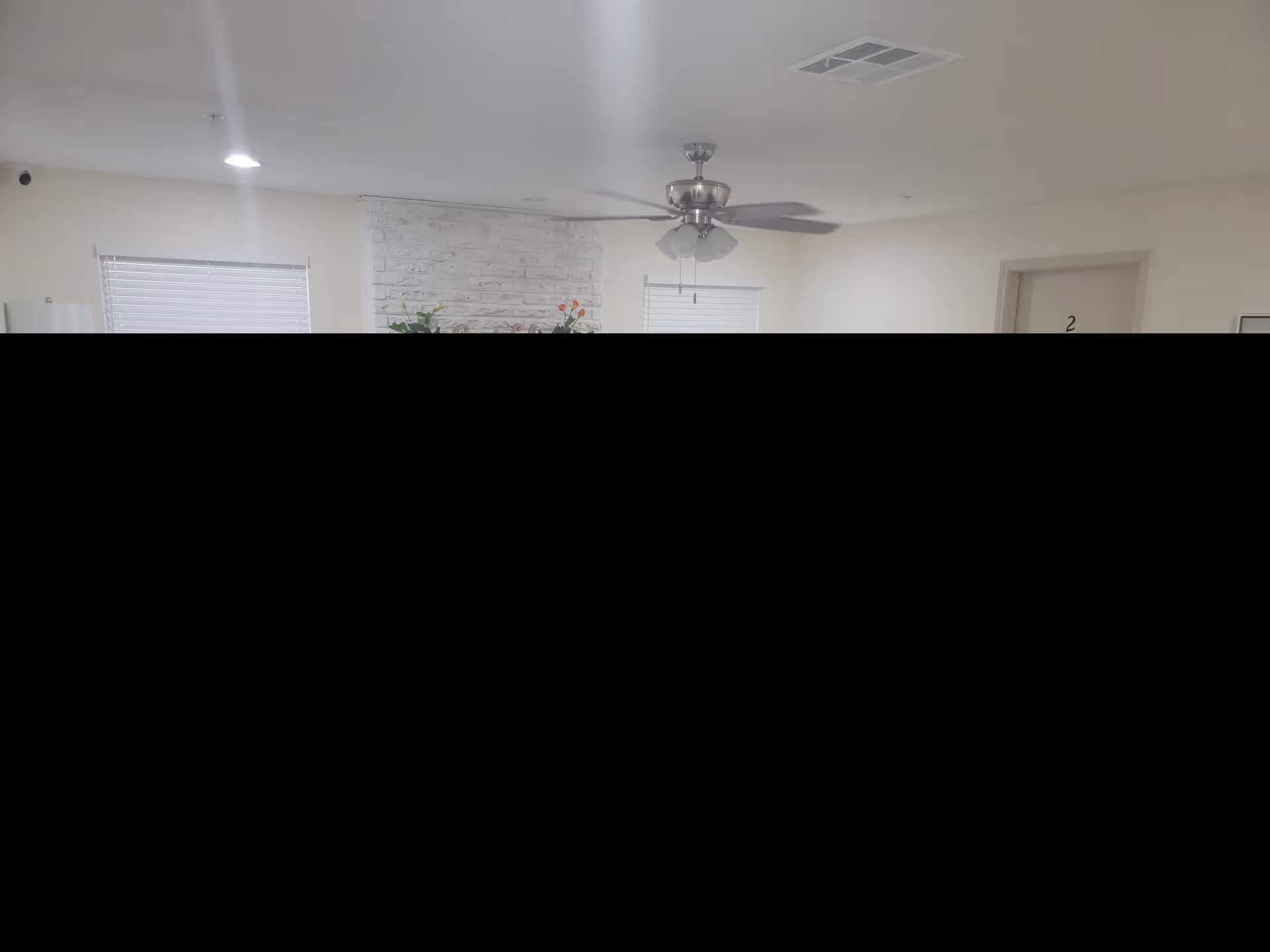 Interior view of a room with a white brick fireplace, two windows with blinds, a ceiling fan with lights, and a door labeled with the number 2. There are plants and decorative items on the fireplace mantel.