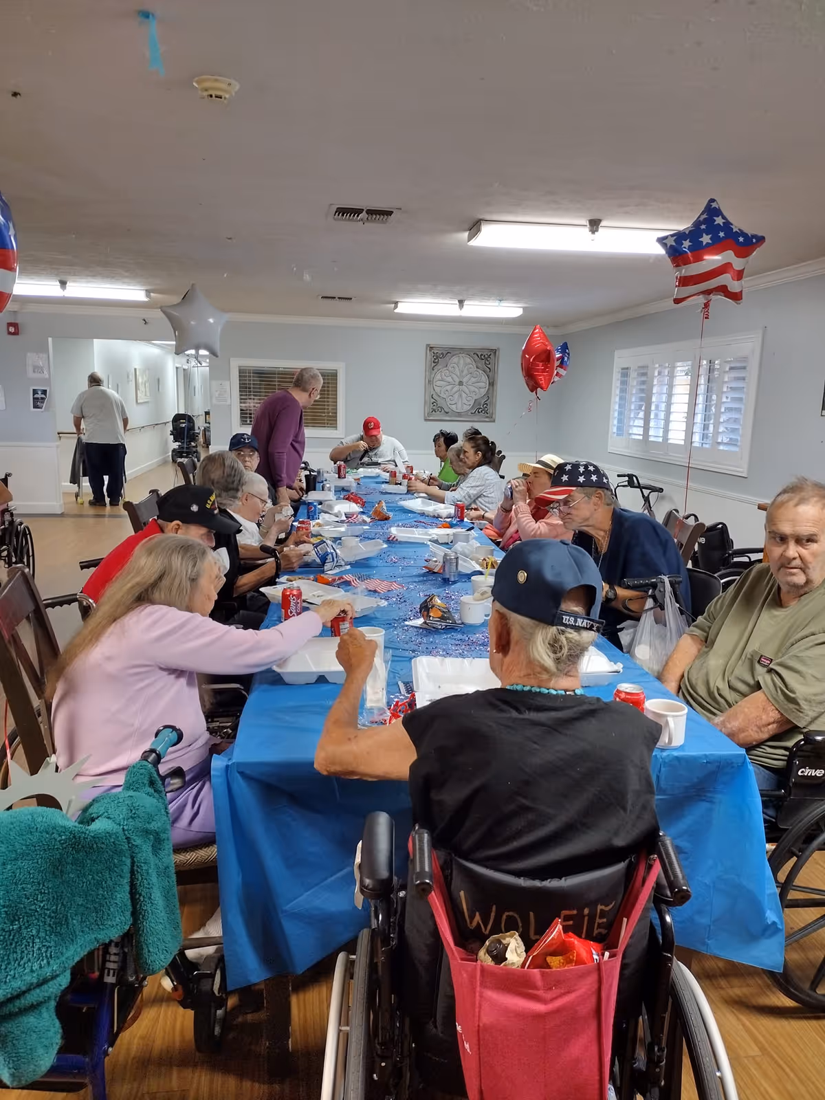 A group of elderly people seated around a long table covered with a blue tablecloth, enjoying a meal together in a communal dining area. The room is decorated with red, white, and blue star-shaped balloons, and some individuals are in wheelchairs. The atmosphere appears social and festive.