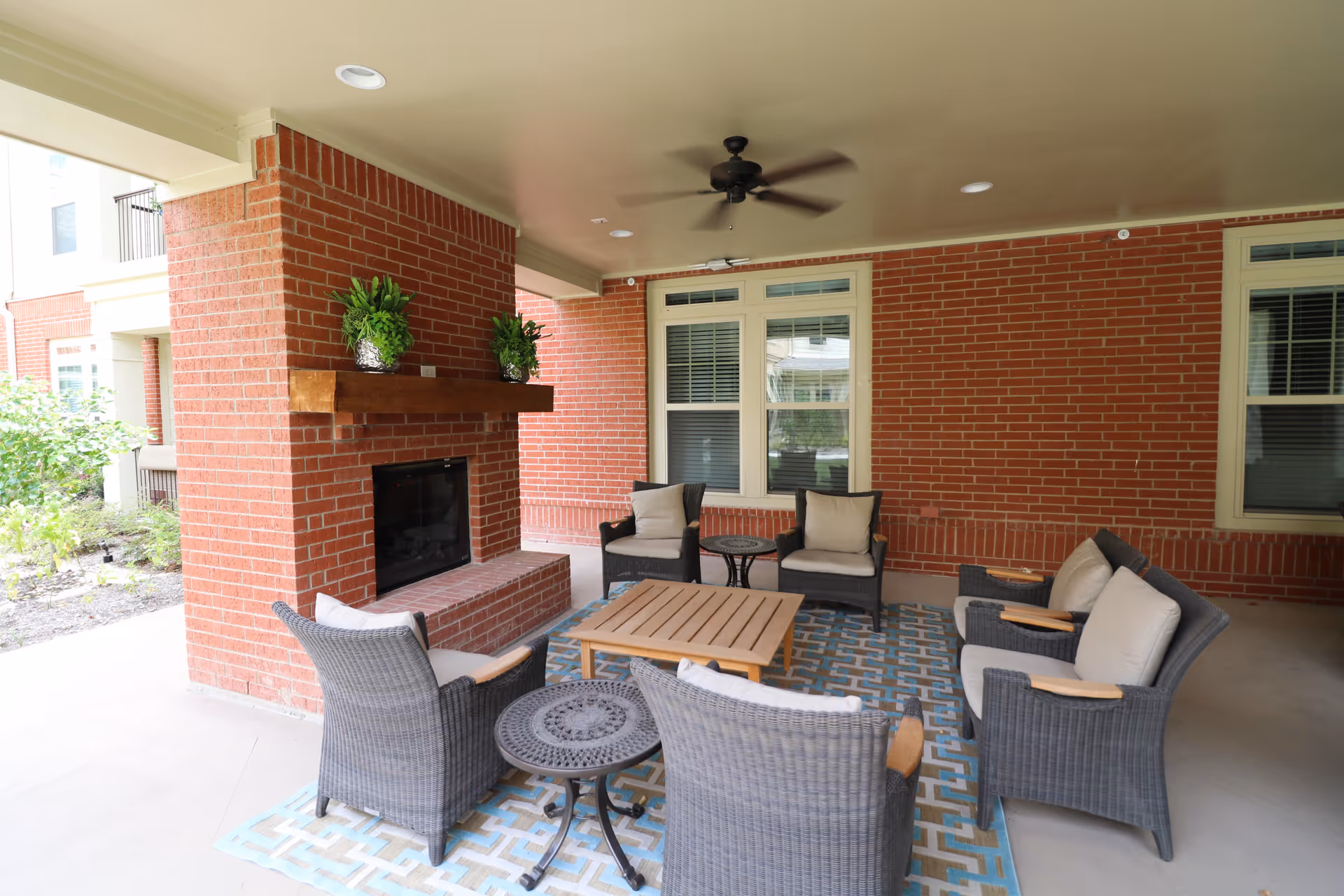 Covered outdoor seating area with a brick fireplace, wicker chairs with cushions arranged around a wooden coffee table on a patterned rug, ceiling fan above, and windows on the brick wall.
