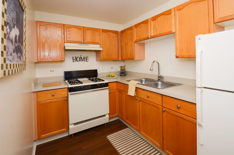A compact kitchen with wooden cabinets, a white gas stove, a double sink with a faucet, a white refrigerator, and a countertop with a bowl of green apples and a beige towel hanging over the sink. There is a framed picture on the left wall and a decorative 'HOME' sign above the stove.
