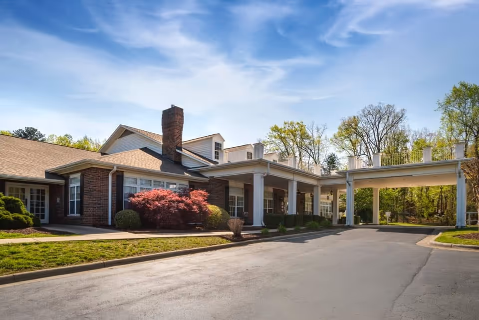 Exterior view of a single-story brick building with a covered entrance supported by white columns. The building is surrounded by green trees and shrubs under a partly cloudy blue sky.