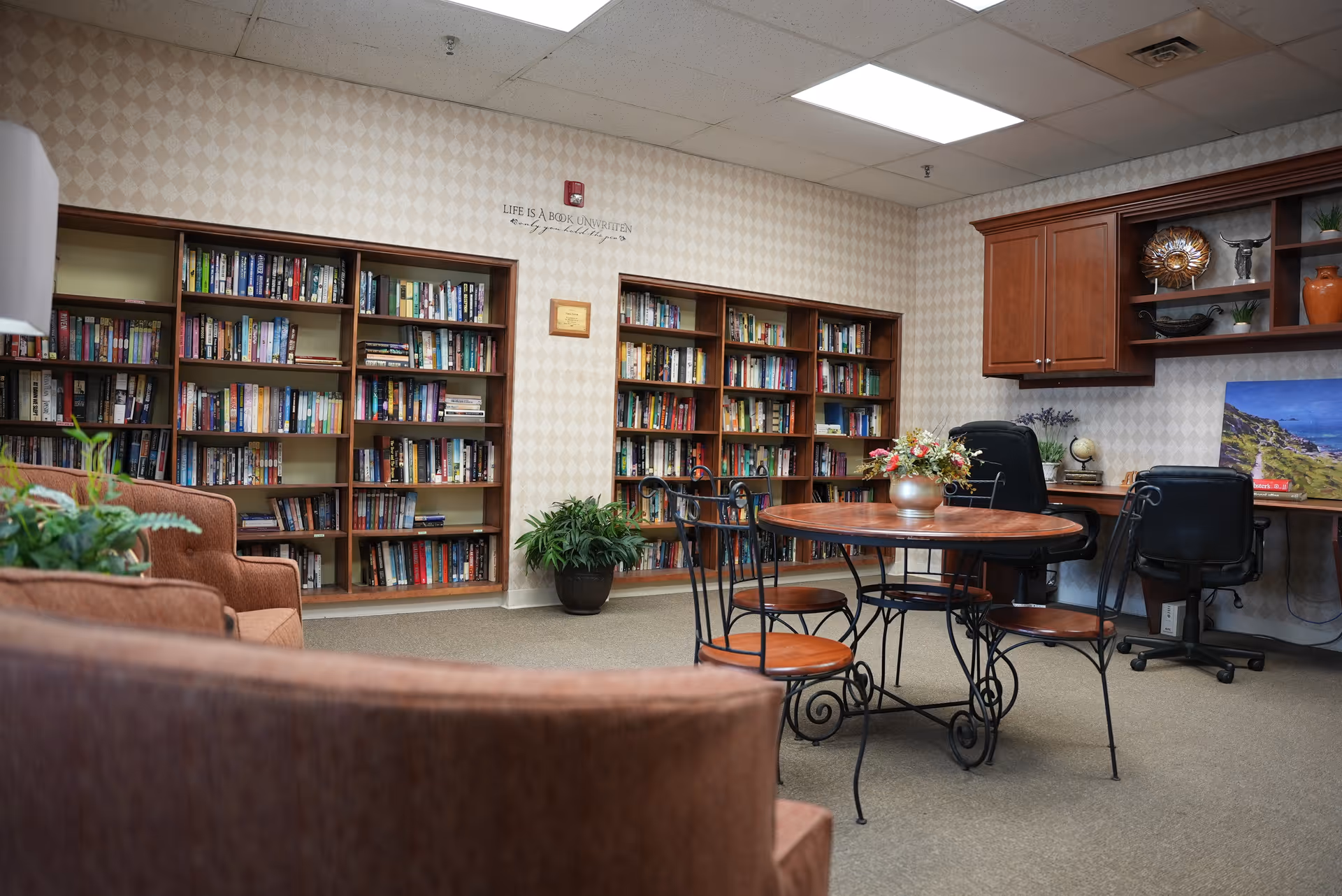 A cozy reading room with multiple bookshelves filled with books, a round wooden table with four metal chairs, two office chairs at a built-in desk with cabinets above, and comfortable upholstered seating in the foreground. The walls have a light patterned wallpaper and there are some plants and decorative items placed around the room.