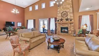 Spacious living room with high ceilings, featuring a stone fireplace with a clock and star decorations above it. The room has multiple seating options including beige sofas and patterned armchairs, a round wooden coffee table, a TV on a stand, and large windows with curtains allowing natural light to fill the space. There are chandeliers hanging from the ceiling and plants placed around the room.