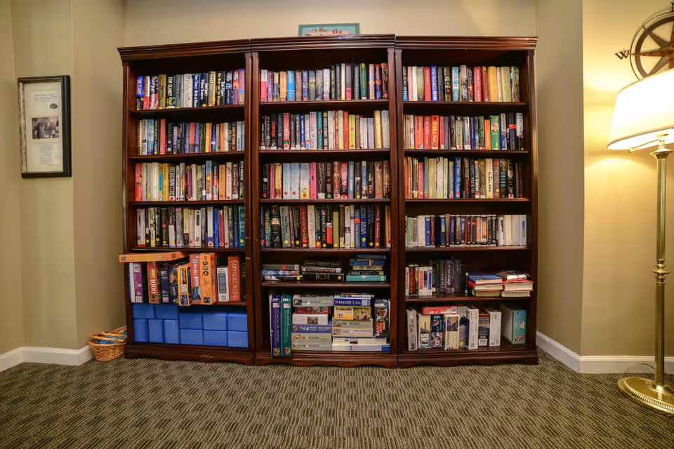 A large wooden bookshelf filled with books, games and boxes against a wall, with a floor lamp to the right.