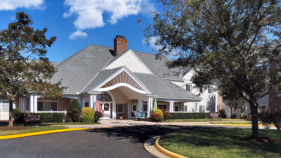 Front exterior view of a senior living facility building with a covered entrance, surrounded by trees, bushes, and a curved driveway under a clear blue sky.