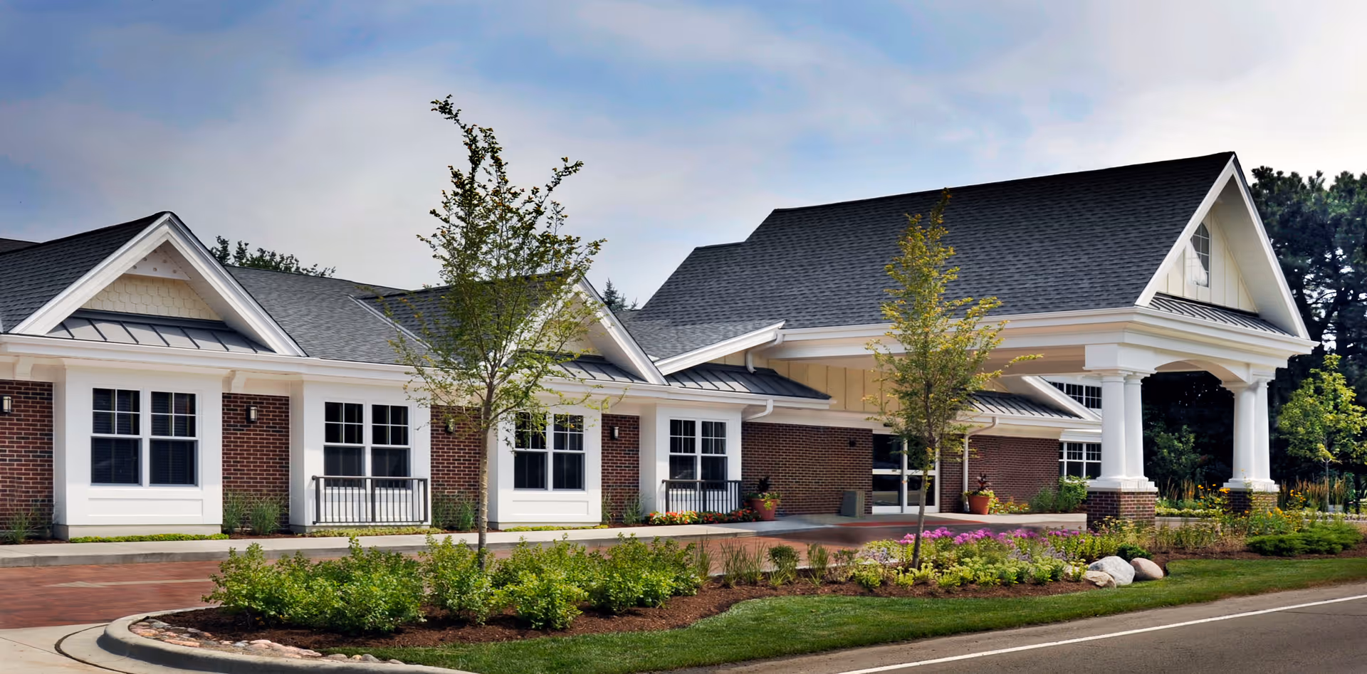 Exterior view of a single-story brick building with white trim and a covered entrance supported by white columns. The building is surrounded by landscaped greenery, small trees, and a paved driveway under a partly cloudy sky.