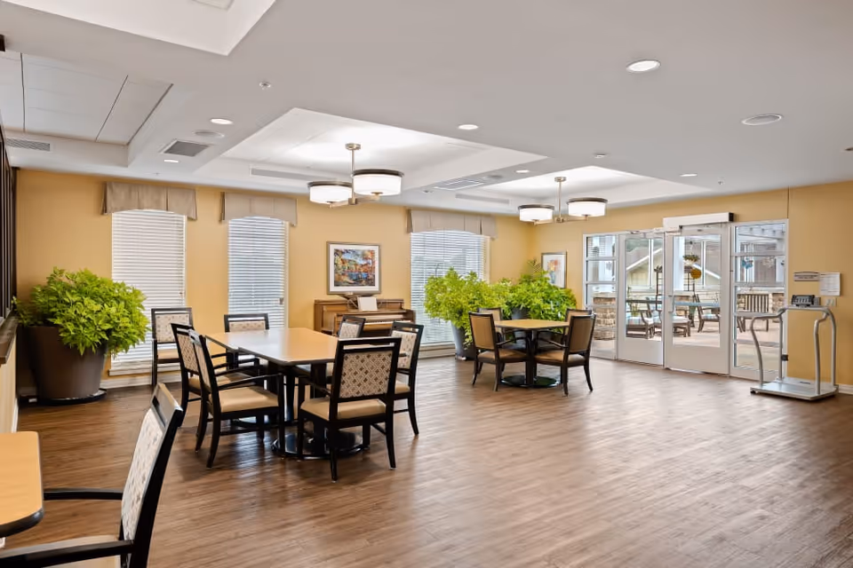 A bright and spacious common area with wooden flooring, beige walls, and large windows with blinds. The room contains two tables with chairs around them, several large green potted plants, a piano against the wall, and glass doors leading to an outdoor patio area with seating. Ceiling lights provide illumination.