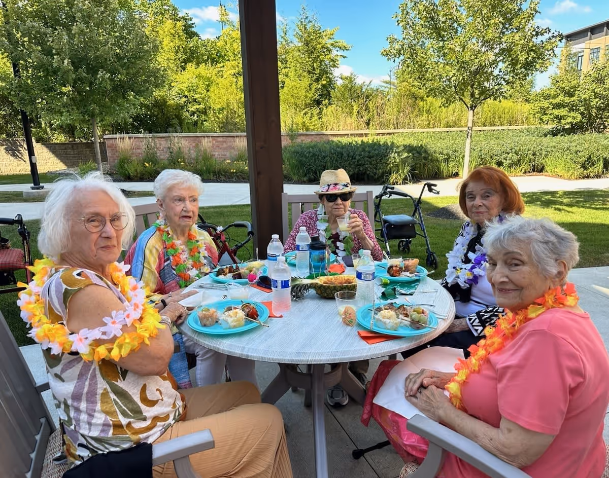 Five elderly women wearing colorful leis are seated around a round outdoor table enjoying a meal together. The table has blue plates with food, water bottles, and a centerpiece made from a hollowed-out pineapple. The setting is a sunny garden area with green grass, trees, and a clear blue sky in the background.
