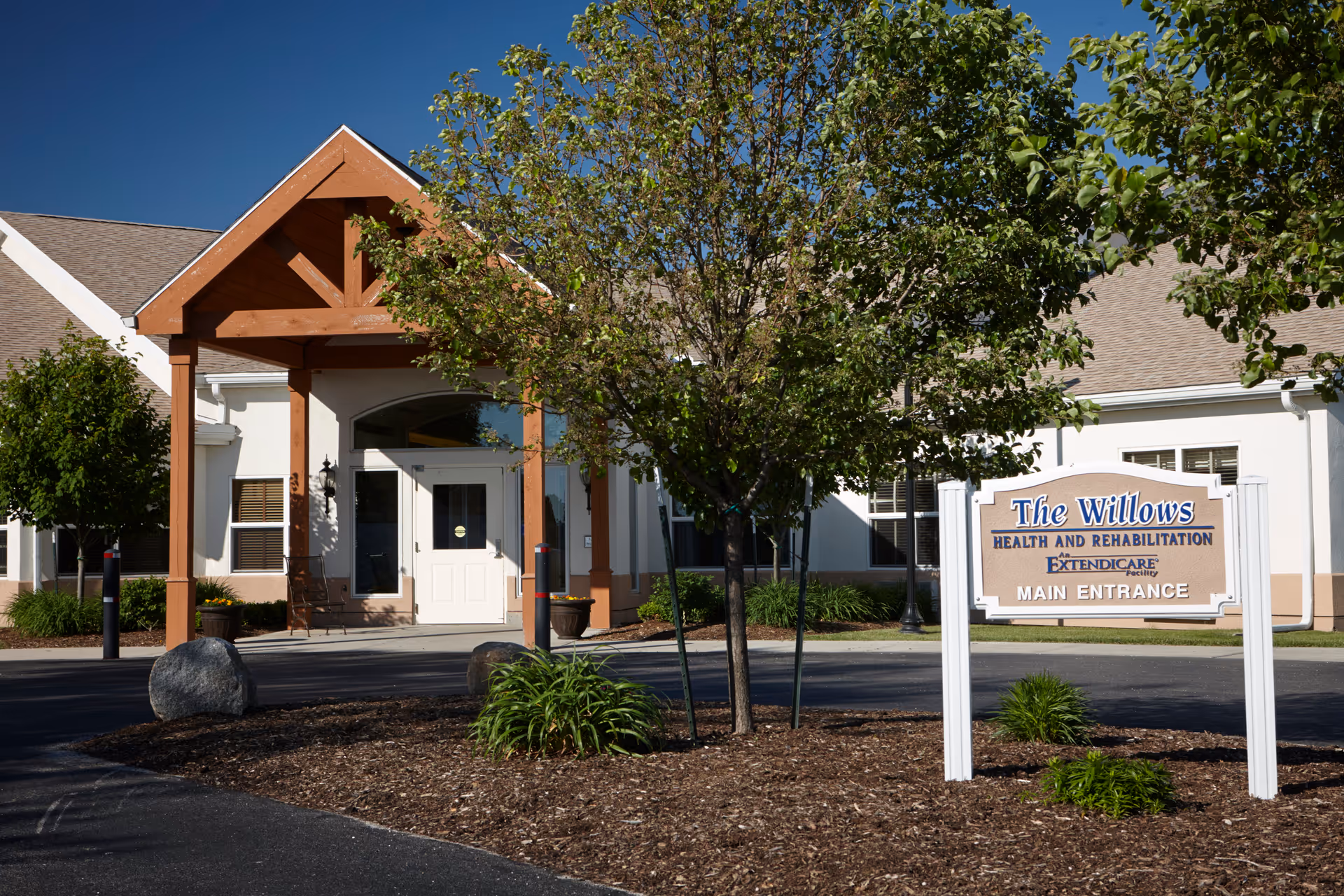 Front entrance of a single-story senior living facility with a covered wooden portico, trees, and a sign reading 'The Willows Main Entrance'.