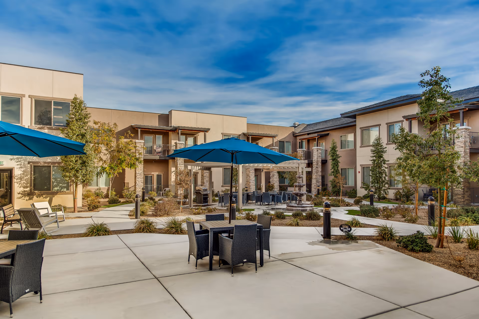 Courtyard of a two-story senior living complex with outdoor seating, blue umbrellas, landscaping, and a central fountain.