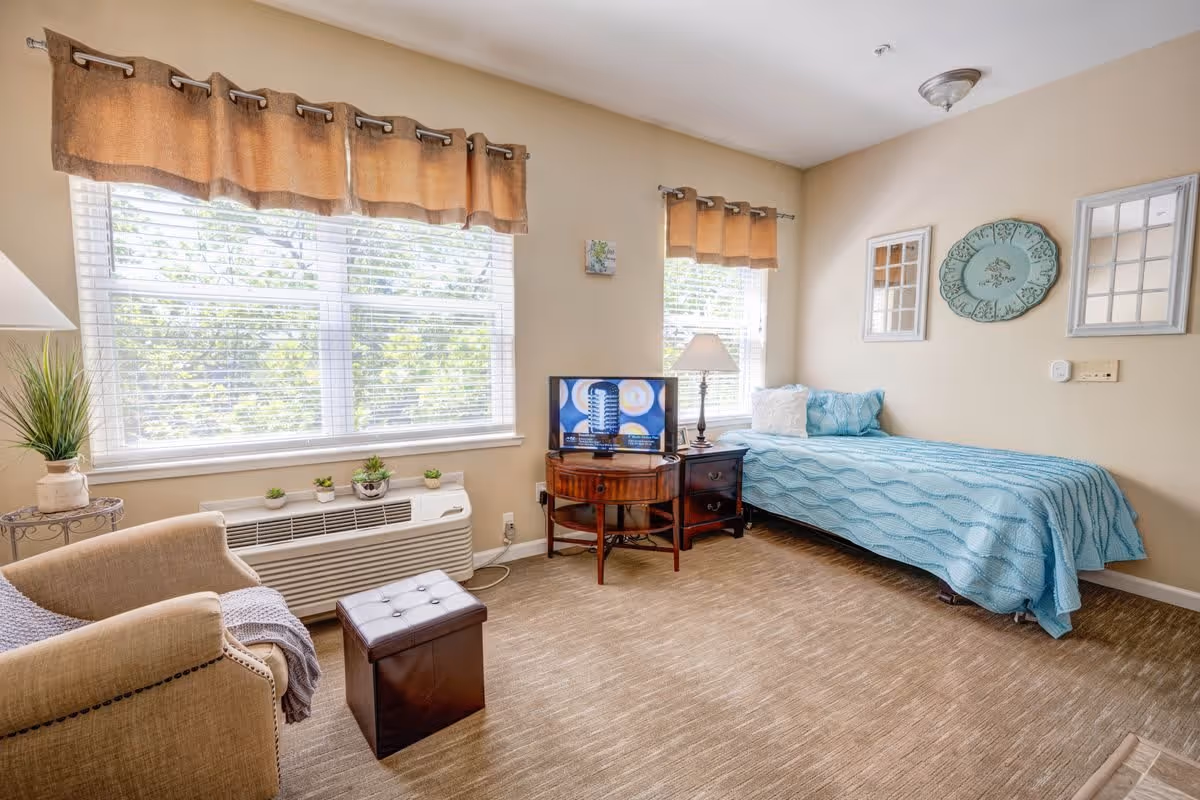A cozy senior living bedroom with a single bed covered in a blue quilt, a small wooden side table with a lamp, a TV on a round wooden table, a beige armchair with a blanket, and large windows with brown valances letting in natural light.