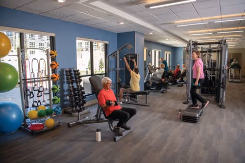 A senior living community fitness room with several elderly individuals exercising using various gym equipment including weights, resistance machines, and step platforms. The room has blue walls, large windows letting in natural light, and a rack of colorful exercise balls and weights.