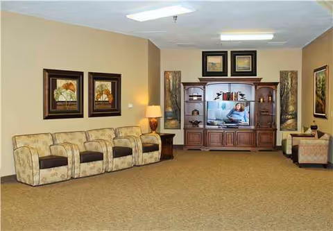 Seniors' living lounge with beige walls, row of patterned chairs, and a wooden entertainment center.