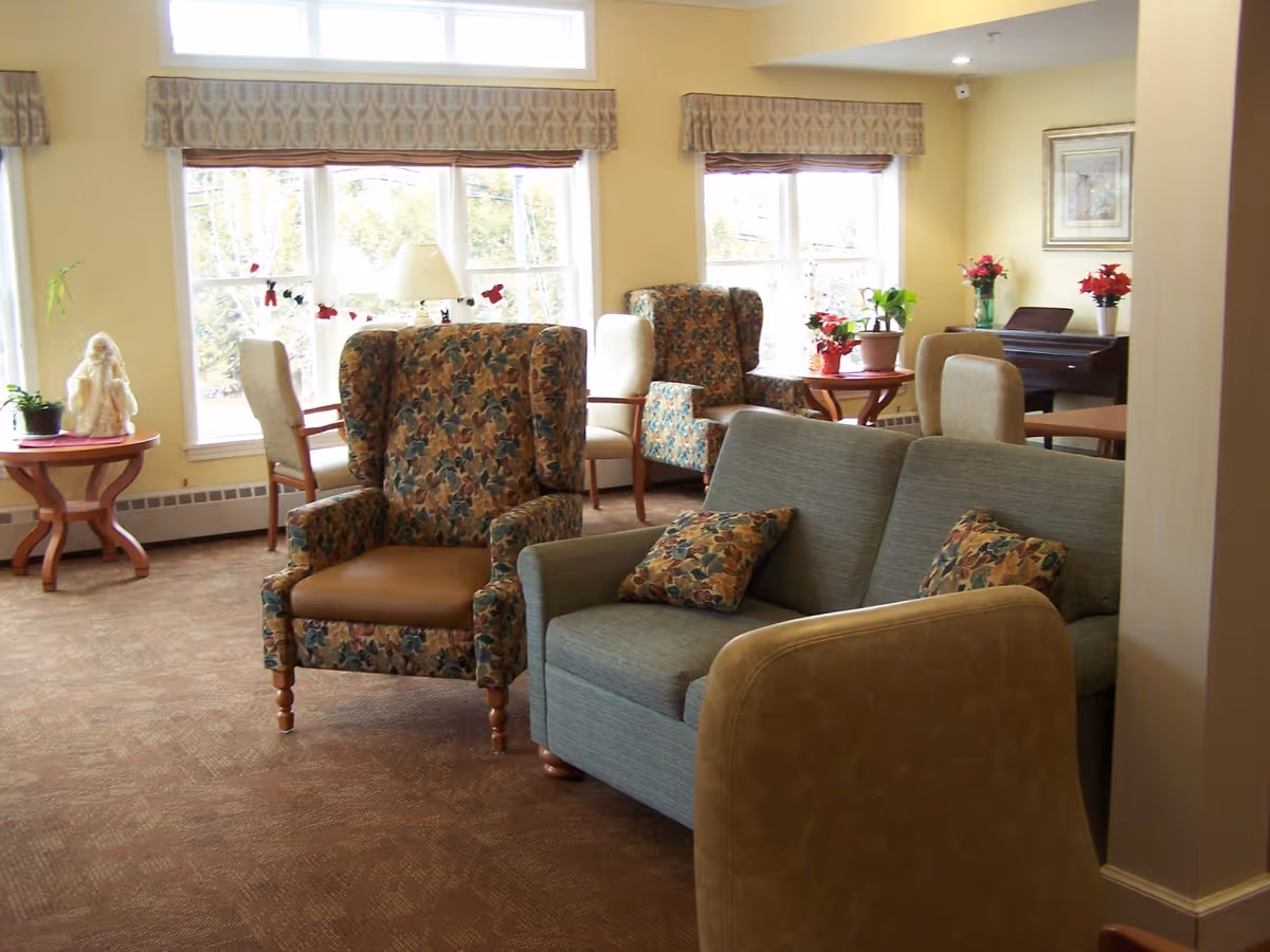 A cozy living room area in Bayview Manor with floral patterned armchairs, a blue loveseat with matching floral pillows, beige chairs, and wooden tables. The room is well-lit with large windows and decorated with potted plants and flowers.