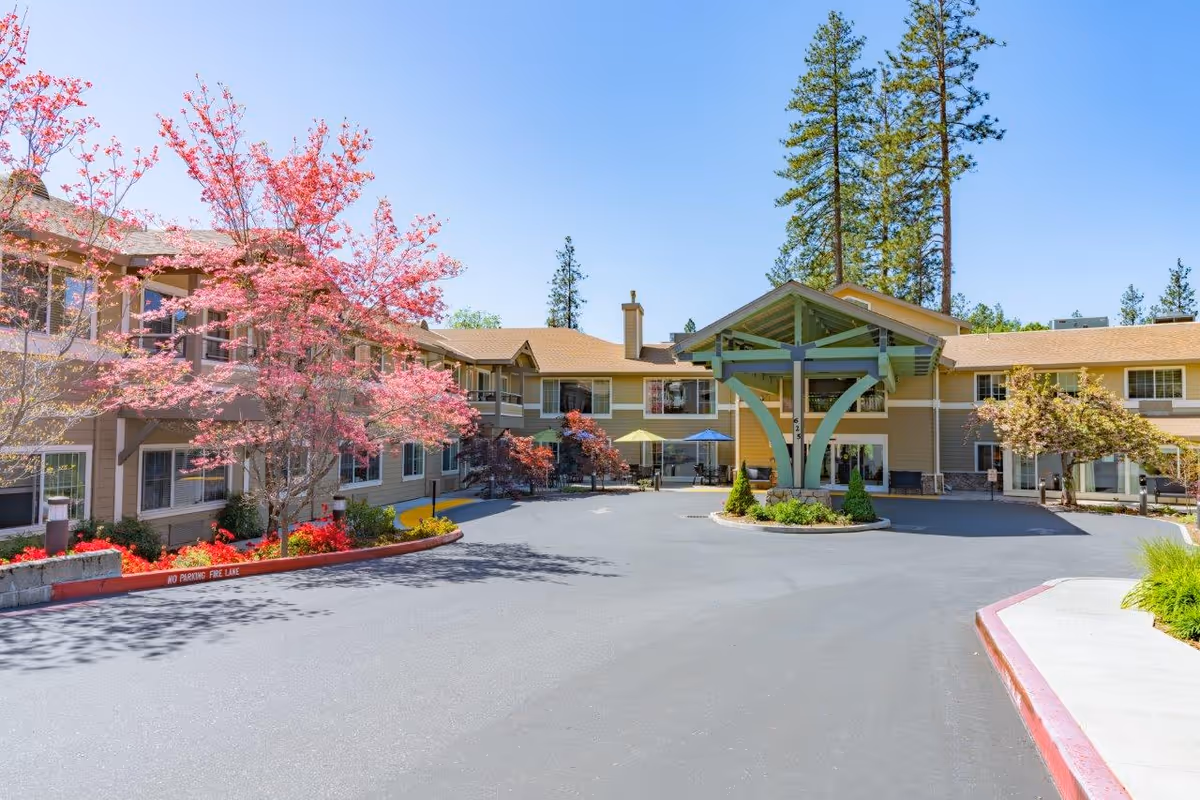 Front entrance of a senior living building with a covered porte-cochère, circular driveway, and flowering trees.
