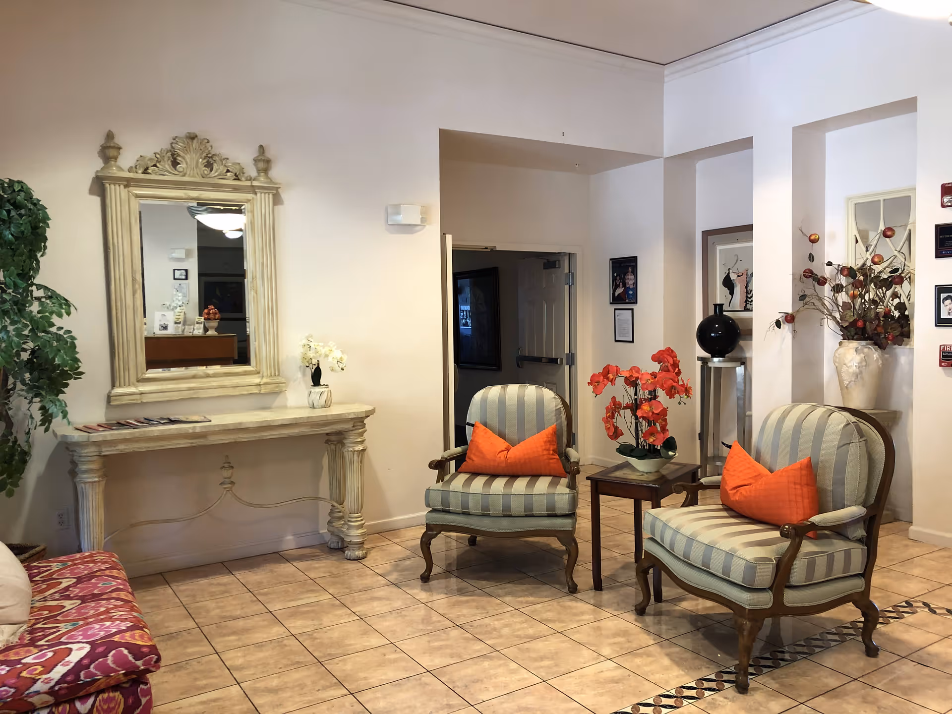 Lobby seating area with two striped armchairs with orange pillows, a small side table with flowers, and a decorative mirror above a console table.