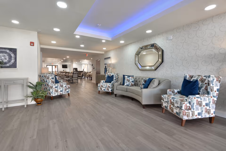 A spacious and well-lit senior living facility common area with modern decor. The room features a light gray curved sofa with blue and patterned cushions, two patterned armchairs, a decorative octagonal mirror on the wall, and a small table with a plant on the left. In the background, there is a dining area with multiple tables and chairs. The ceiling has recessed lighting and a blue LED accent light.