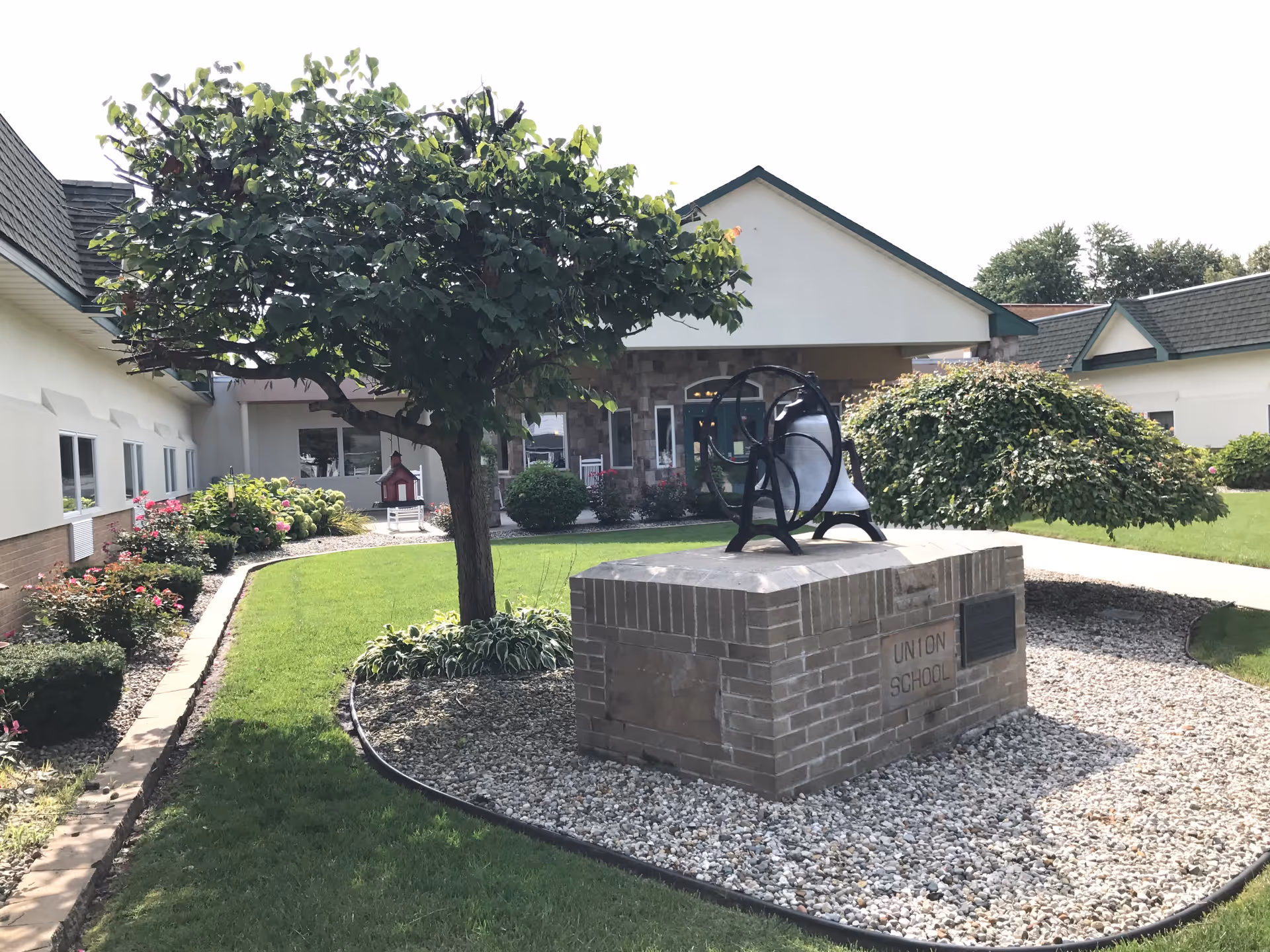 Outdoor courtyard area of Union Court Assisted Living featuring a large tree, well-maintained green lawn, flower beds, and a brick pedestal with a mounted bell labeled 'Union School'. The building with white walls and green roof trim surrounds the courtyard.