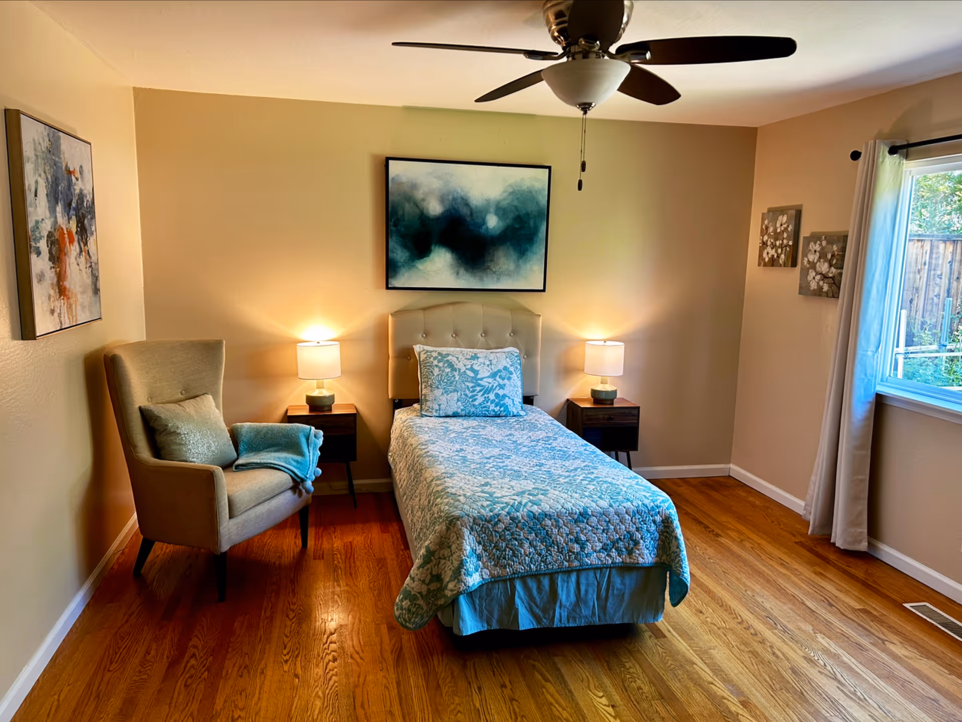 A cozy bedroom with a single bed covered in a blue and white patterned quilt and matching pillow. On either side of the bed are small wooden nightstands with lamps. A beige upholstered armchair with a blue throw blanket and pillow is positioned to the left. The room has hardwood floors, beige walls, a ceiling fan, and a window with white curtains letting in natural light. Abstract and floral artwork decorate the walls.