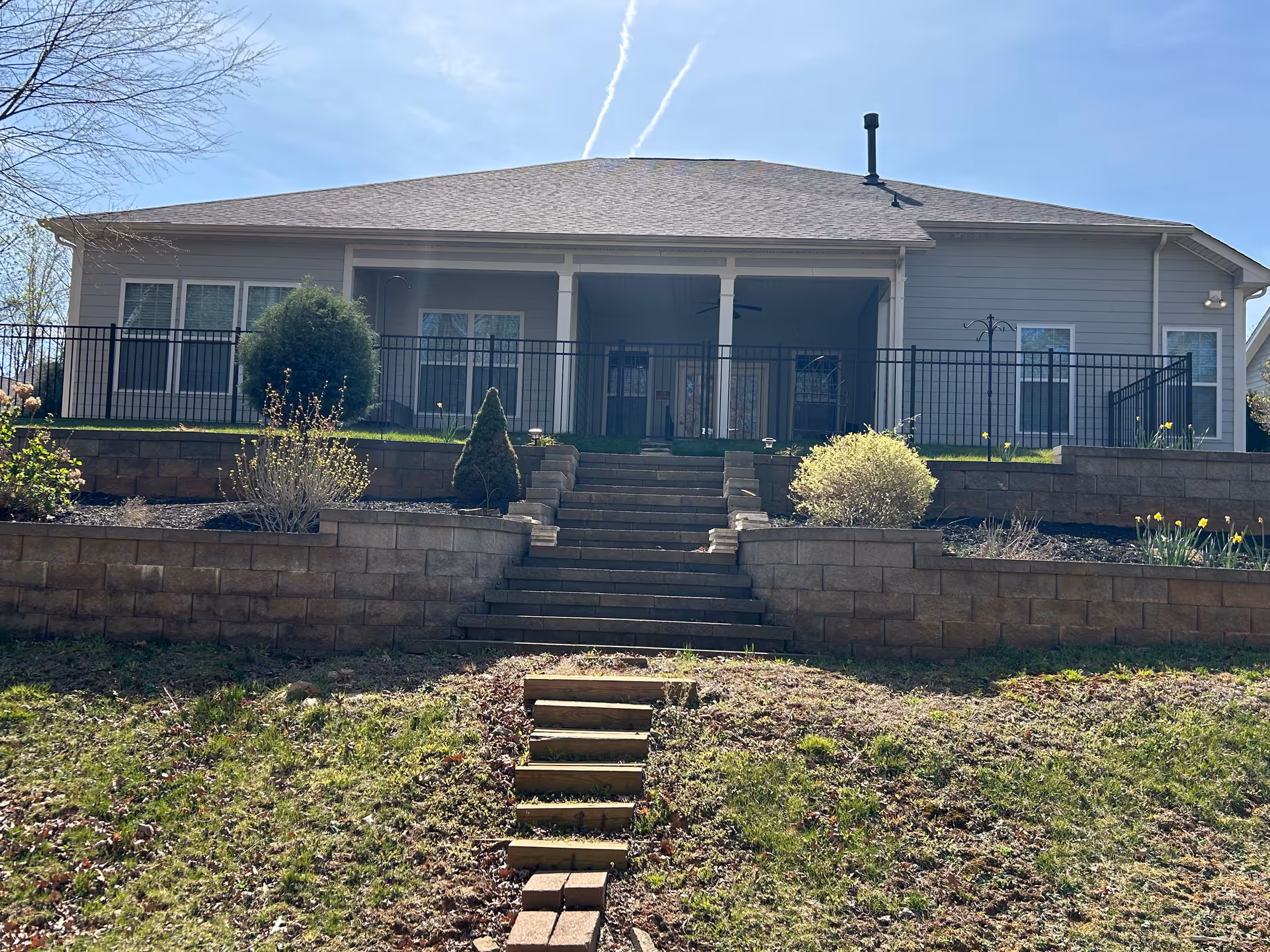 Exterior view of a single-story senior living facility building with a sloped roof, a covered porch area, and multiple windows. The building is elevated with stone retaining walls and stairs leading up to the entrance. There are various shrubs and plants in the landscaped area around the stairs.