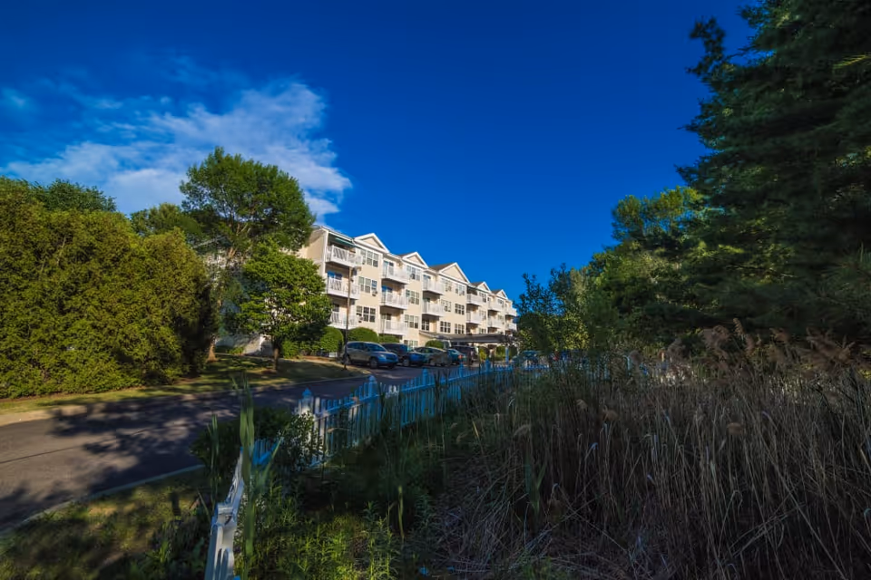 Multi-story senior living apartment building with balconies, parked cars, trees and a white fence under a clear blue sky.