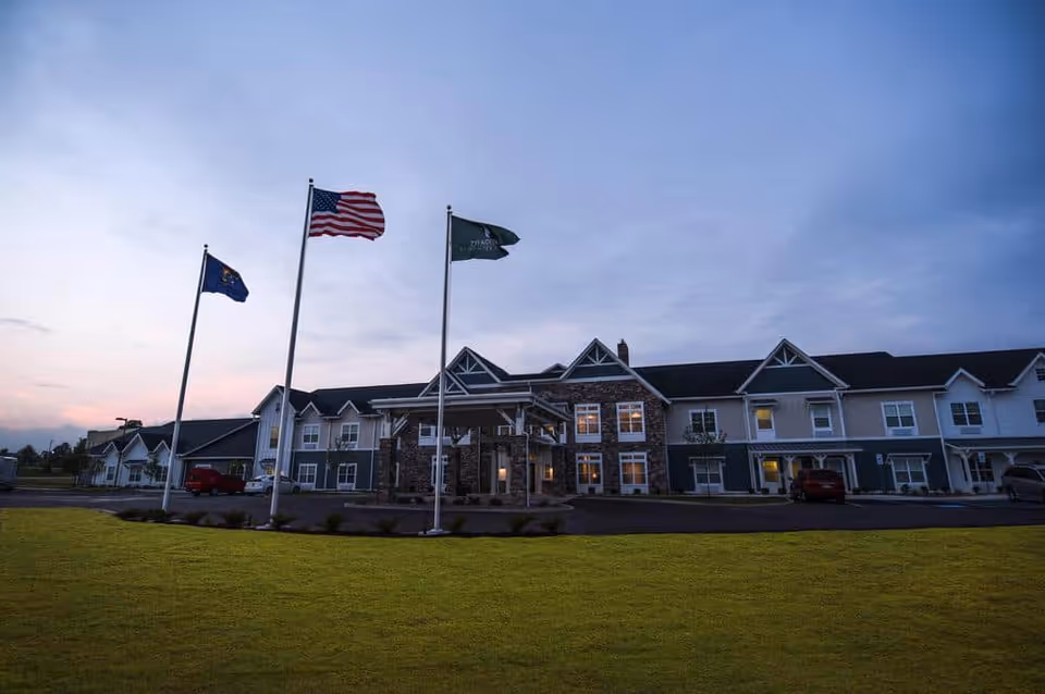 Exterior view of Traditions at Hunter Station facility at dusk with three flagpoles displaying flags in front of a two-story building with a stone and siding facade and a well-maintained lawn.