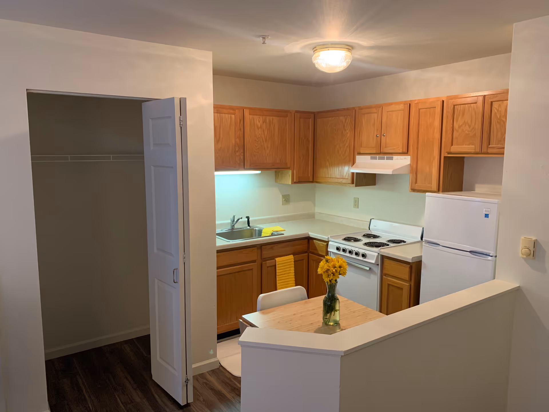 A small kitchen area with wooden cabinets, a white refrigerator, a white stove with an overhead vent, and a sink. There is a small table with a vase of yellow flowers and a white chair. To the left, there is an open closet with a white folding door. The floor is dark wood, and the walls are light-colored.