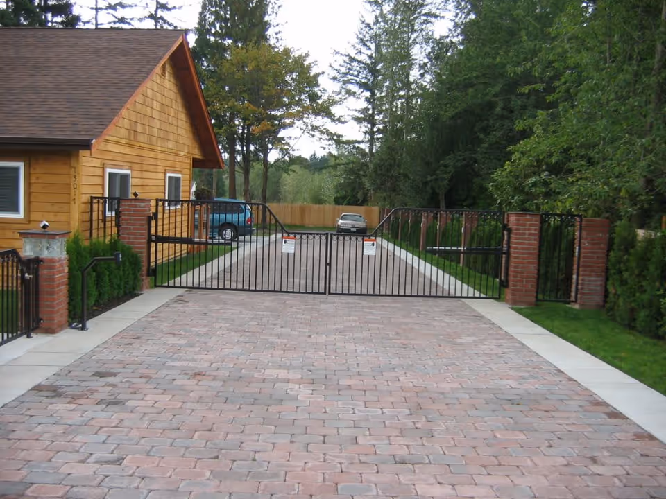 Brick-paved driveway leading to a closed black metal gate in front of a wood-sided house and trees.
