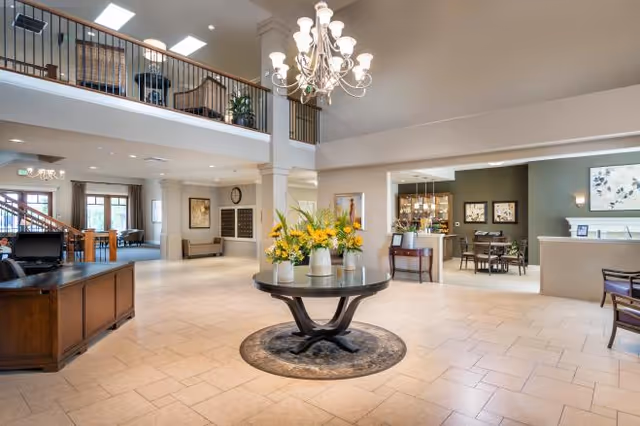 Spacious senior living facility lobby with a central round table of flowers, chandelier, seating areas, and an upstairs balcony.