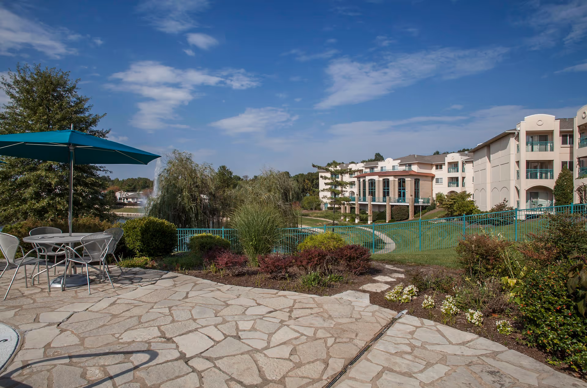 Outdoor patio area with a stone floor, metal table and chairs under a teal umbrella. In the background, there is a landscaped garden with bushes, trees, and a walking path leading to a multi-story building under a partly cloudy blue sky.